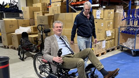 Finanzstaatssekretär Dr. Worms sitzt in einem Velobike und testet es in der Fertigungshalle, neben ihm Alexander Kraft, Pressesprecher des Unternehmens.